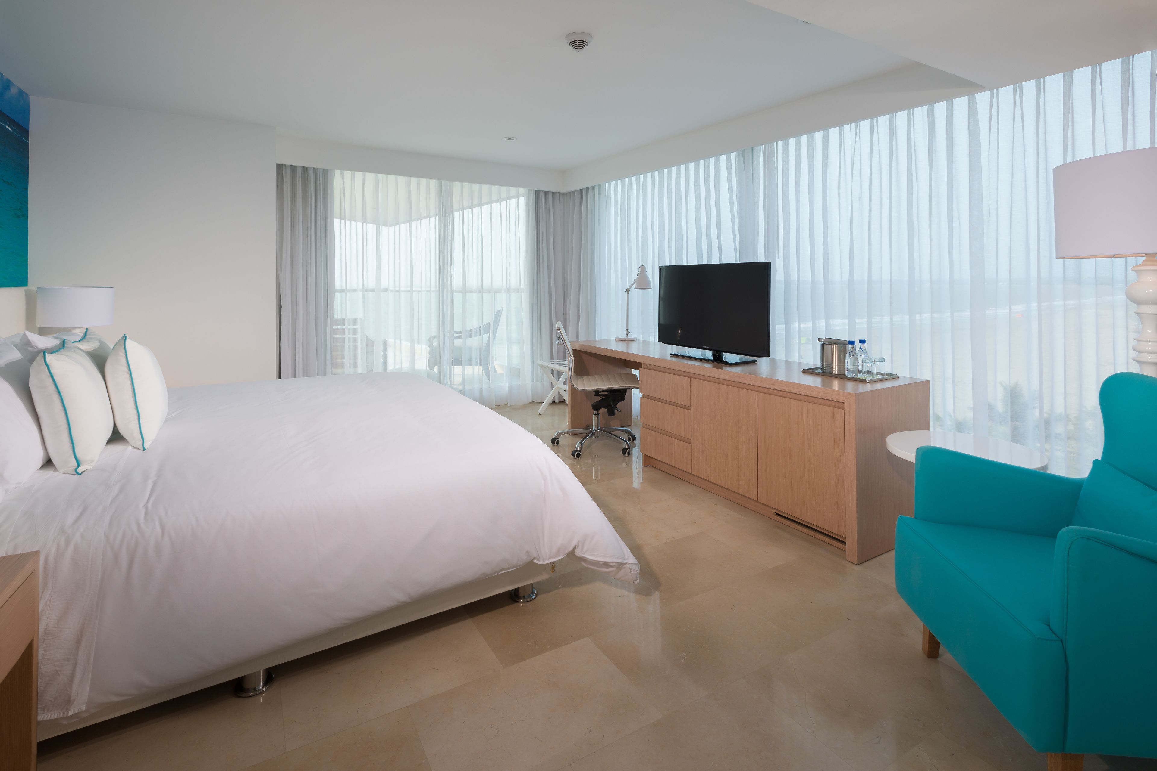 A hotel room with a large bed, a desk with a TV, a chair, and a view of the city through sheer curtains.
