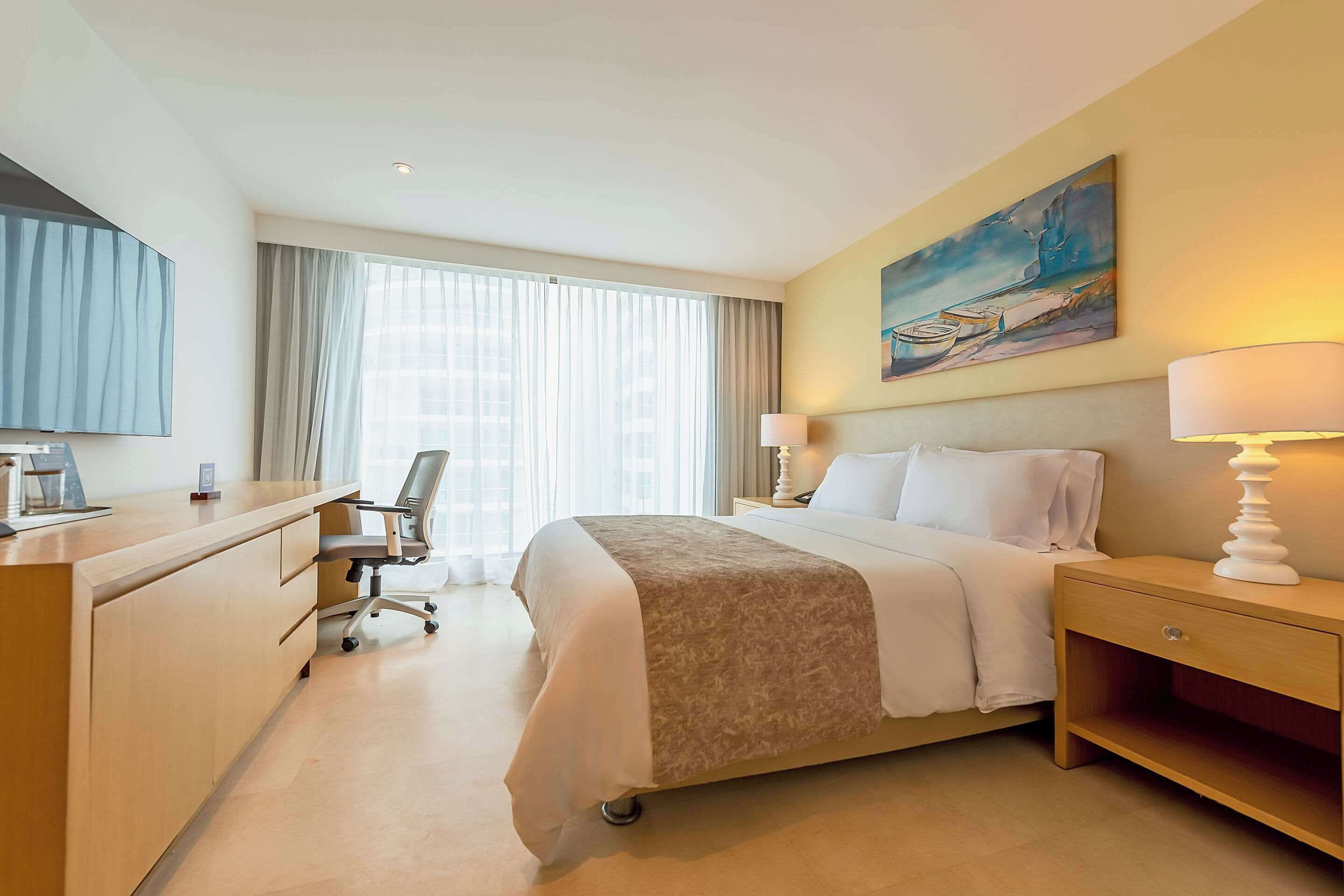 A hotel room with a bed, desk, chair, and a painting on the wall.