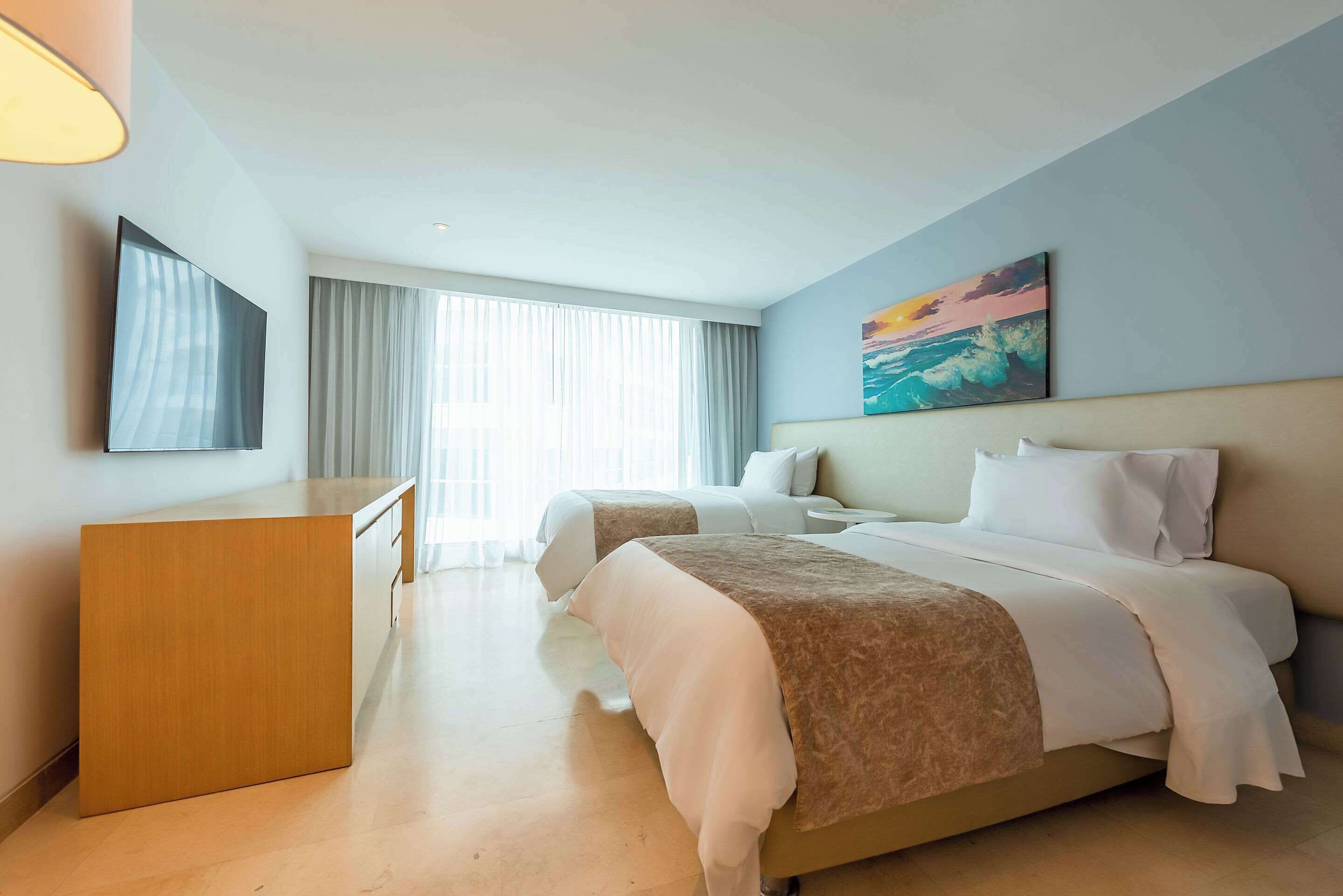 A hotel room with a desk, two beds, a painting, and a large window with curtains.
