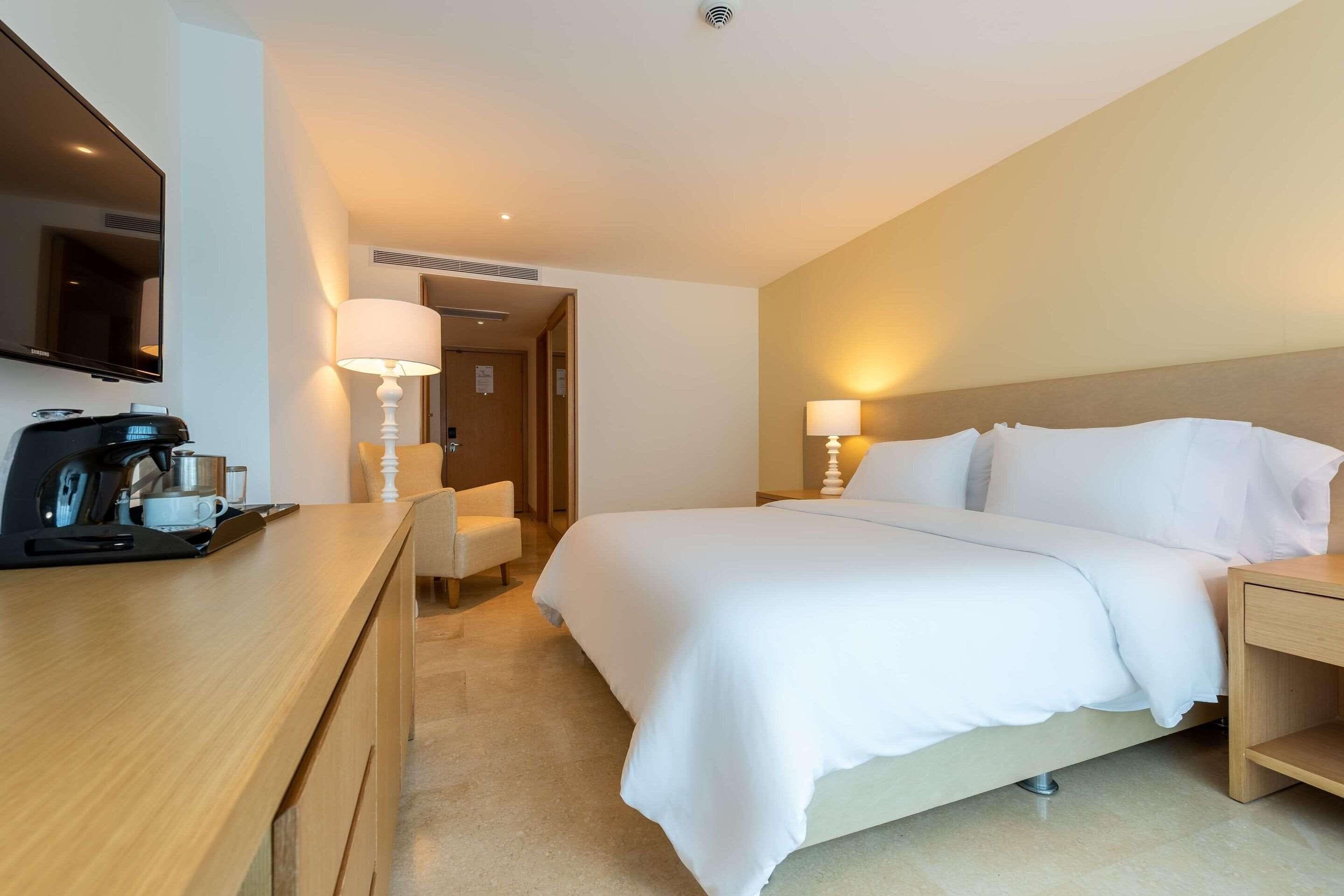 Una habitación de hotel con cama, mesitas de noche, escritorio con cafetera, silla y televisor en la pared.