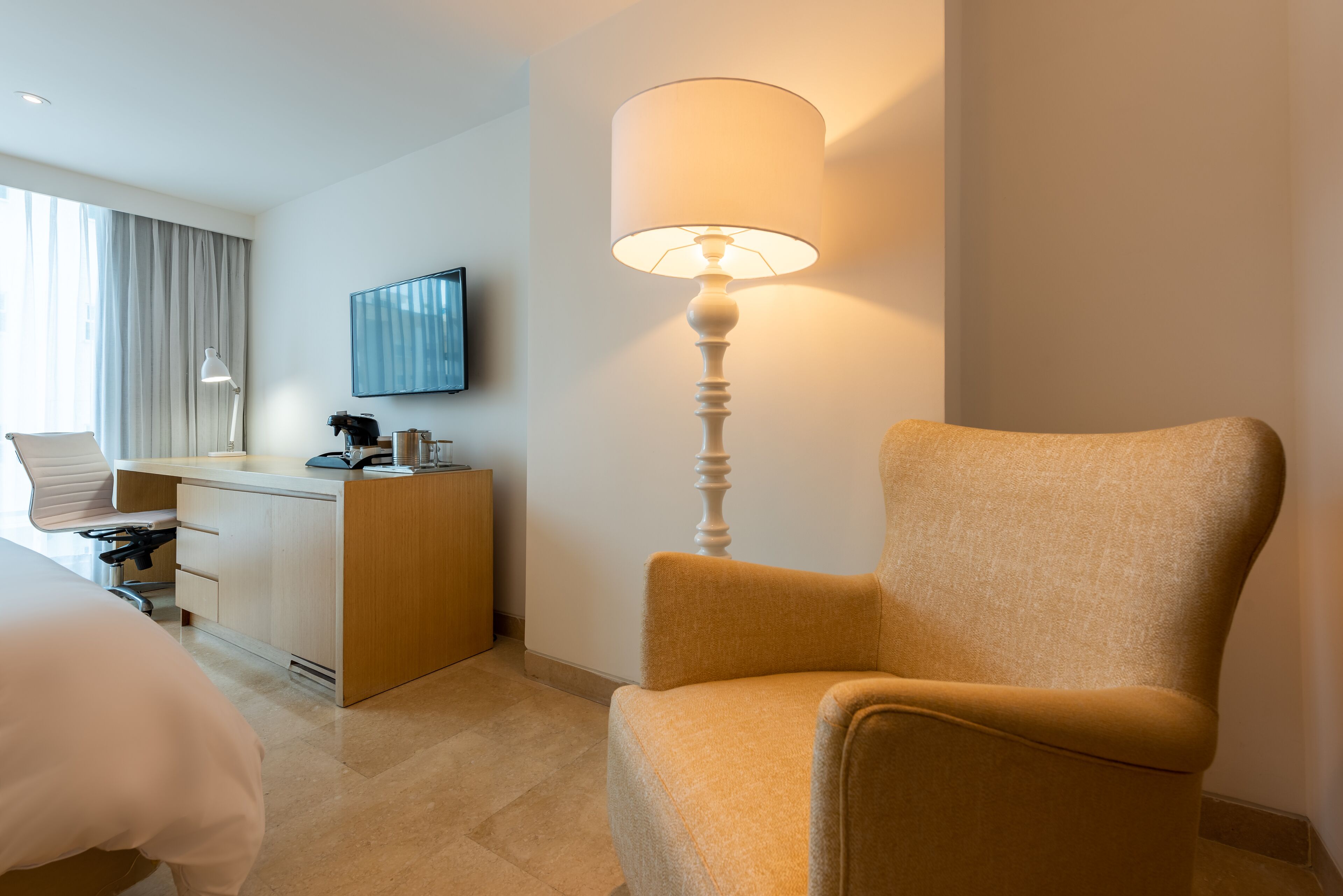A hotel room with a bed, a desk with a lamp, a chair, and a television on the wall.