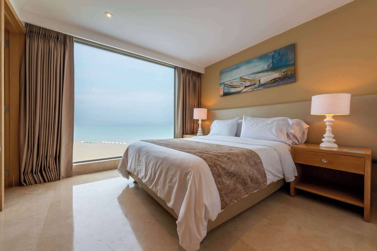 Une chambre d’hôtel avec un grand lit, une table de chevet, un tableau au mur et une vue sur la plage par la fenêtre.