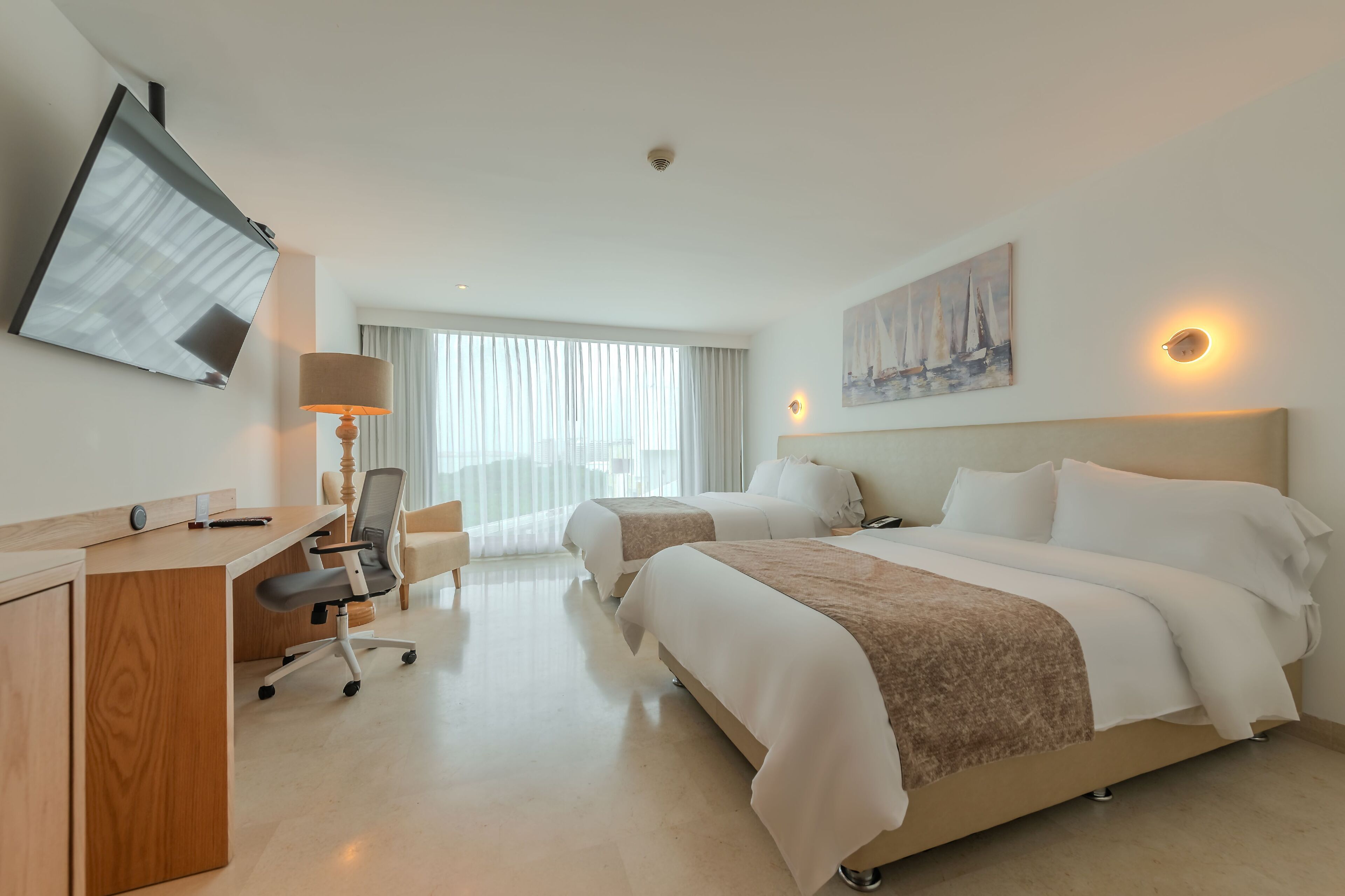 A hotel room with a desk, two beds, a TV, a painting, and a window with curtains.