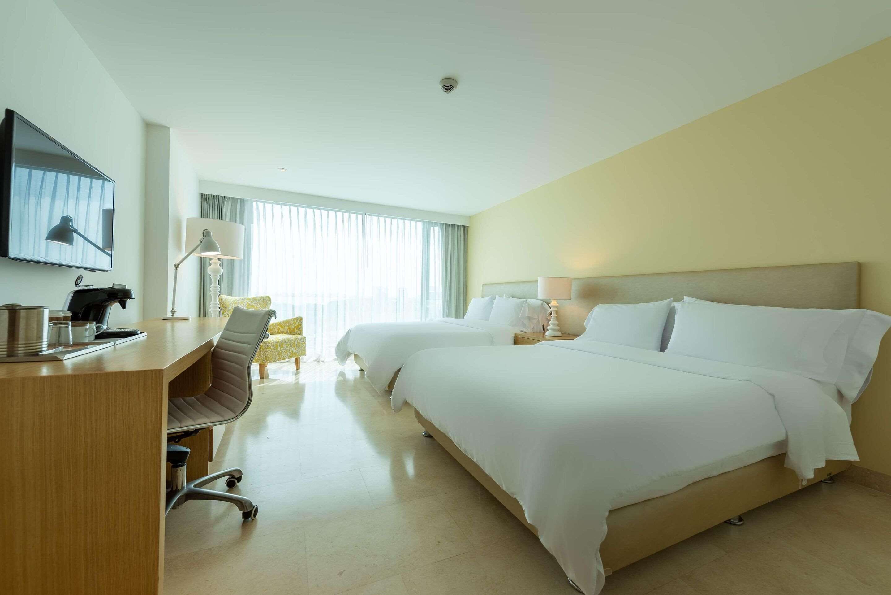 A hotel room with two beds, a desk, a chair, a TV, and a window with curtains.