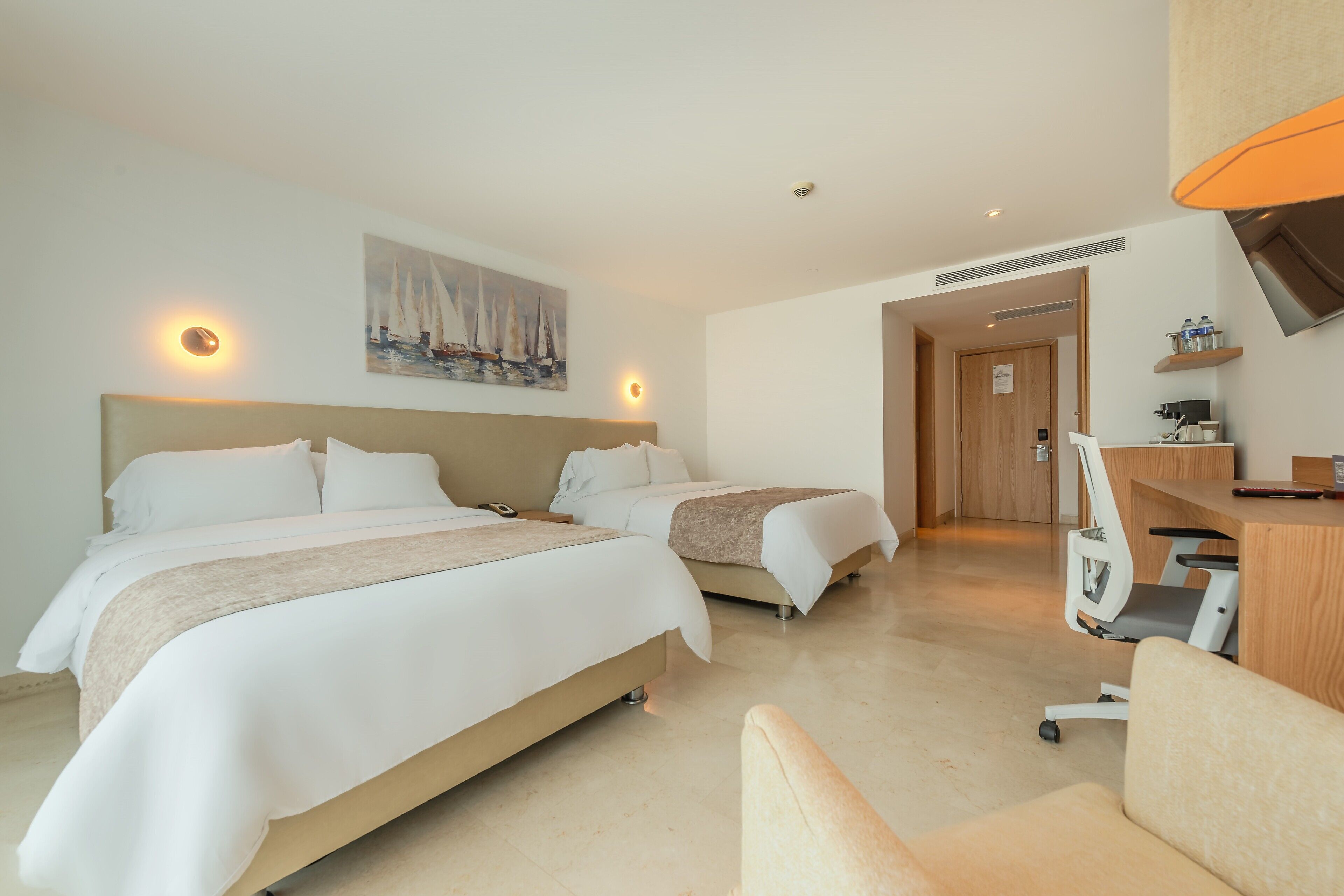 A hotel room with two beds, a desk, a chair, a painting on the wall, and a wooden door leading to another room.