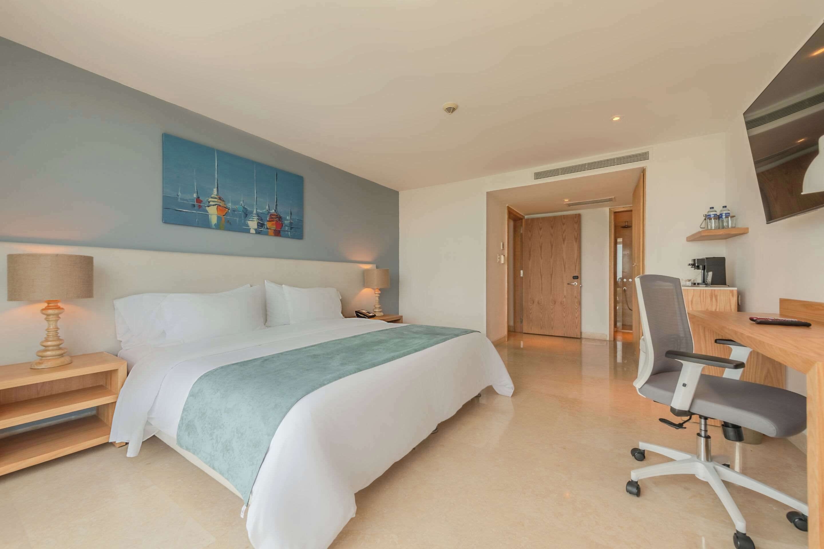 A hotel room with a bed, bedside table, lamp, desk, chair, and a painting on the wall.