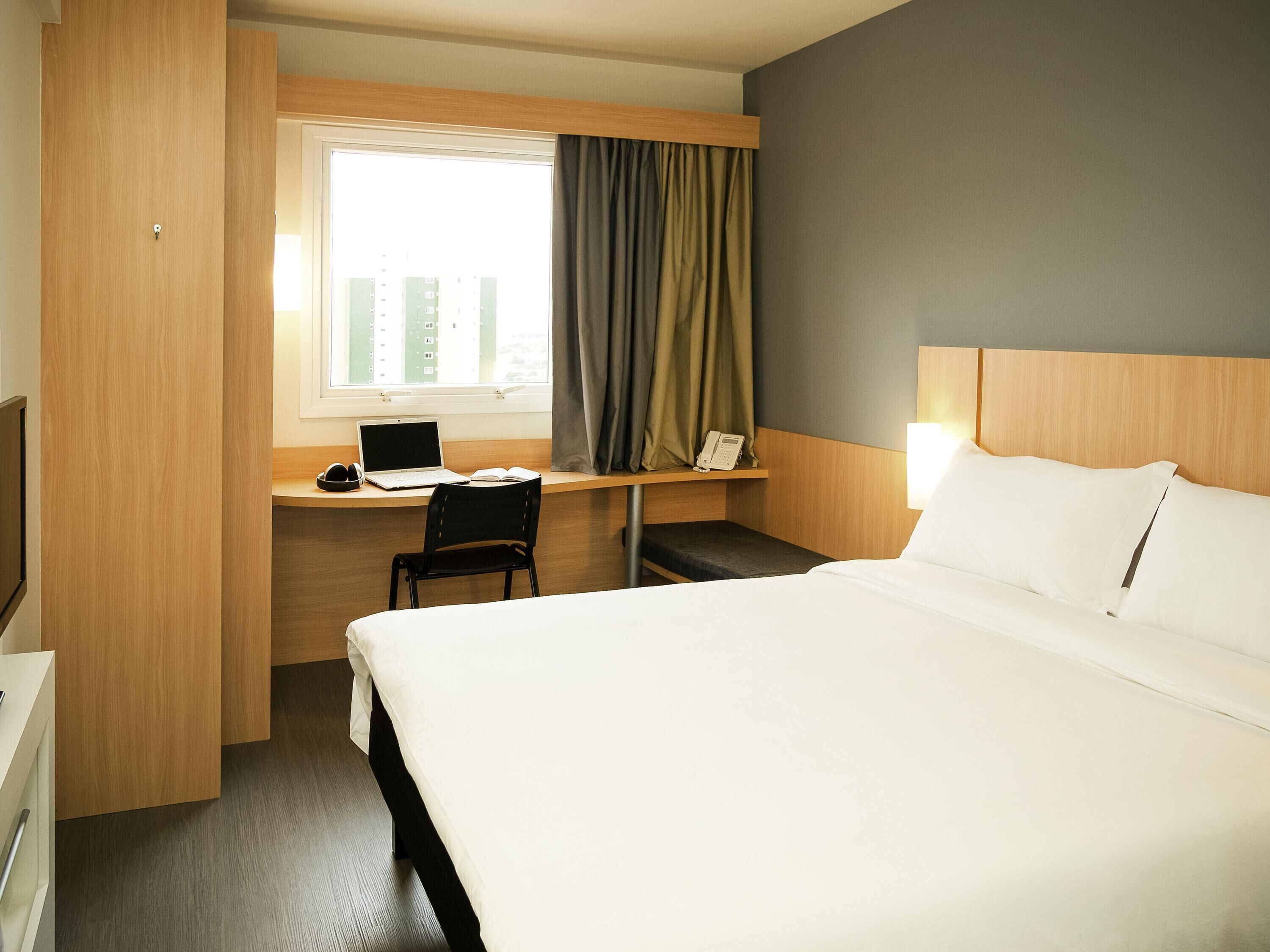 A hotel room with a bed, a desk with a laptop, a chair, a bedside table with a phone, and a window with curtains.