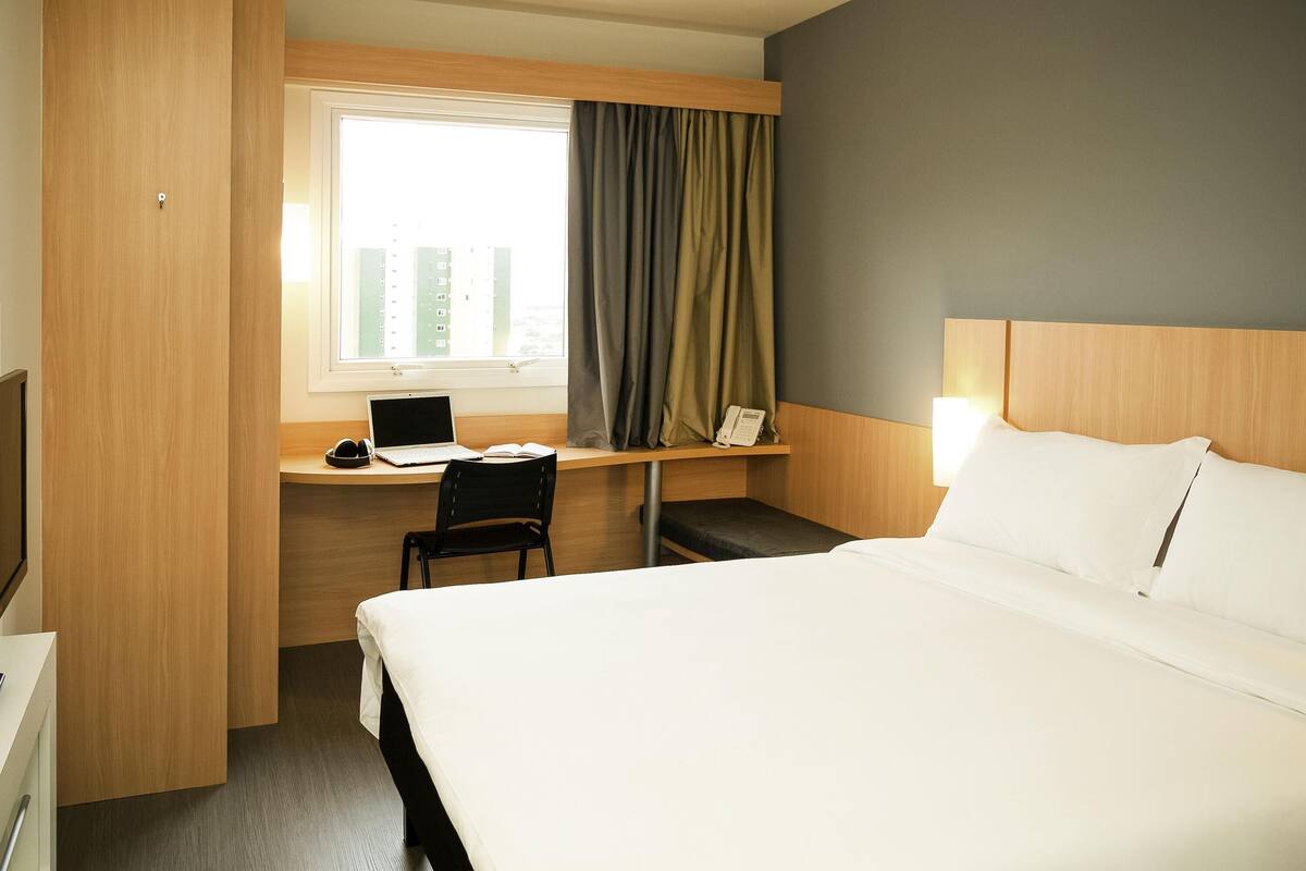 A hotel room with a bed, a desk with a laptop, a chair, a bedside table with a phone, and a window with curtains.