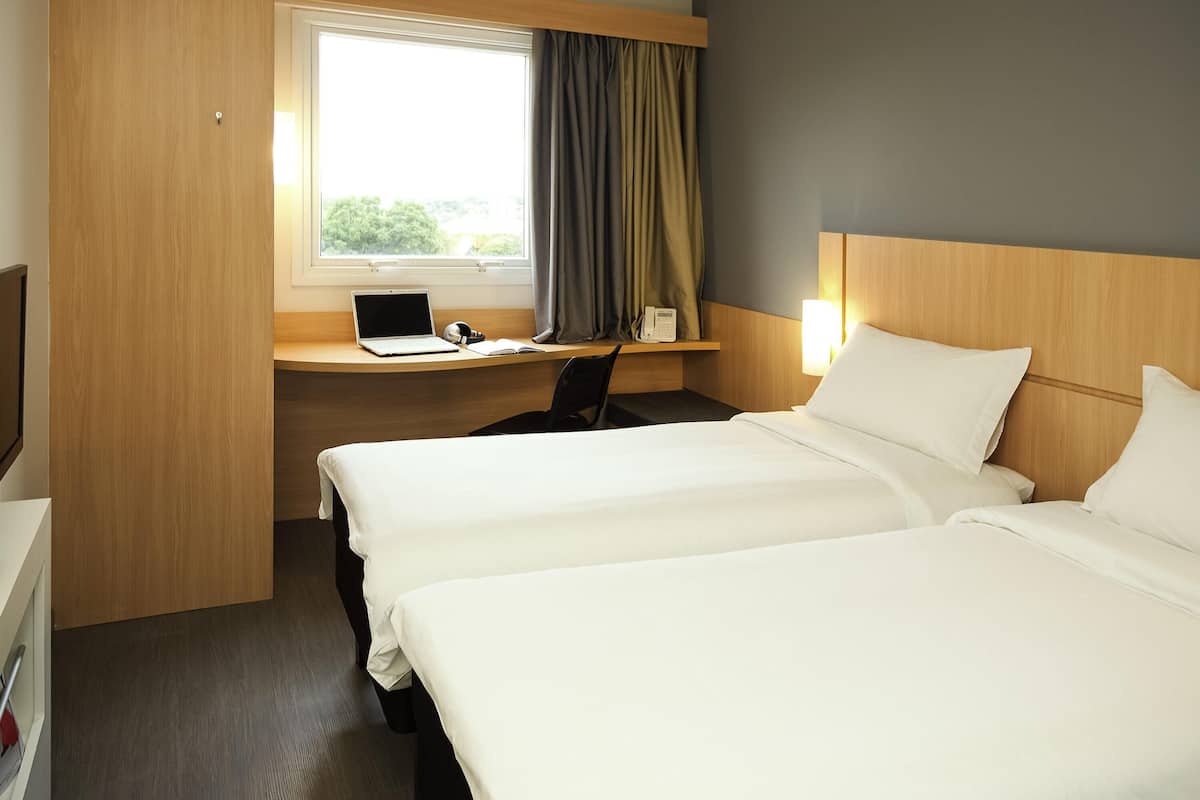 A hotel room with two beds, a desk with a laptop, a chair, and a window with curtains.
