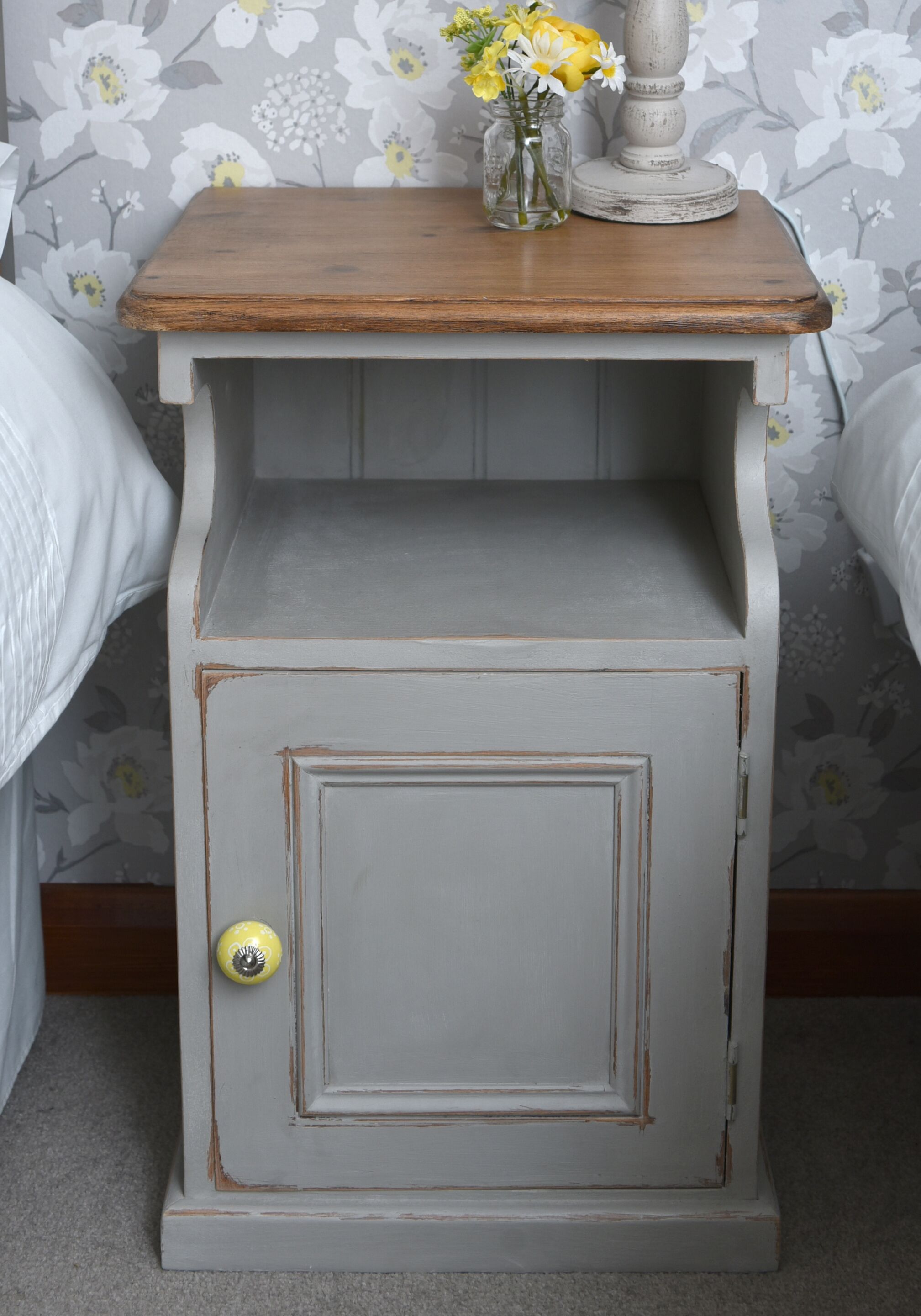A small wooden nightstand with a drawer, a vase with flowers, and a candle.