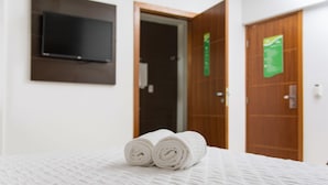 Una cama bien tendida, toallas blancas enrolladas, un televisor de pantalla plana montado en la pared y dos puertas de madera al fondo.