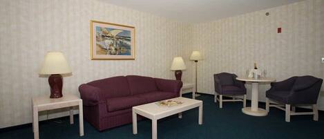 A hotel room with a purple sofa, two armchairs, a small table with a magazine, two lamps, and a painting on the wall.