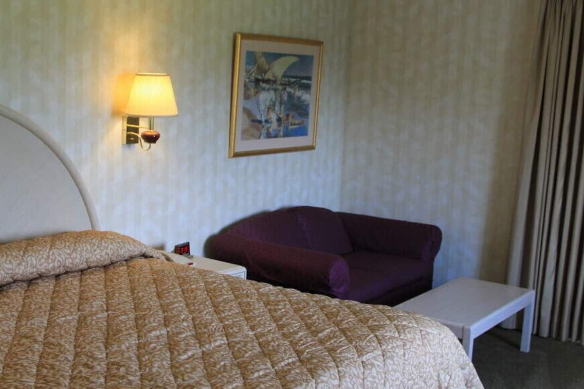 A hotel room with a bed, a chair, a small table, a wall-mounted lamp, and a painting on the wall.