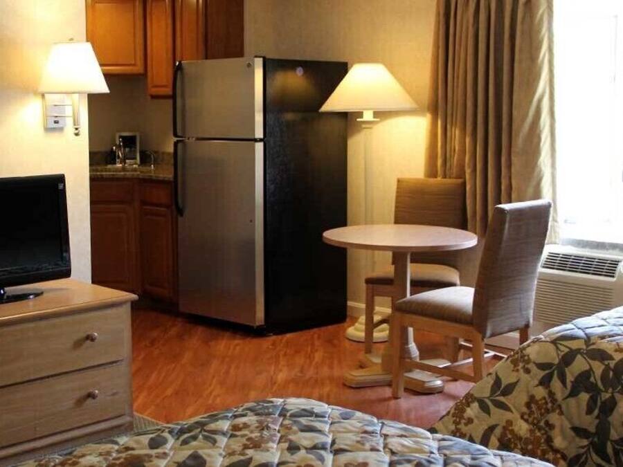 Habitación de hotel con cama, escritorio con computadora, refrigerador, silla, mesita y ventana con cortinas.