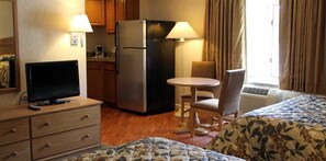 A hotel room with a bed, a desk with a computer, a refrigerator, a chair, a small table, and a window with curtains.