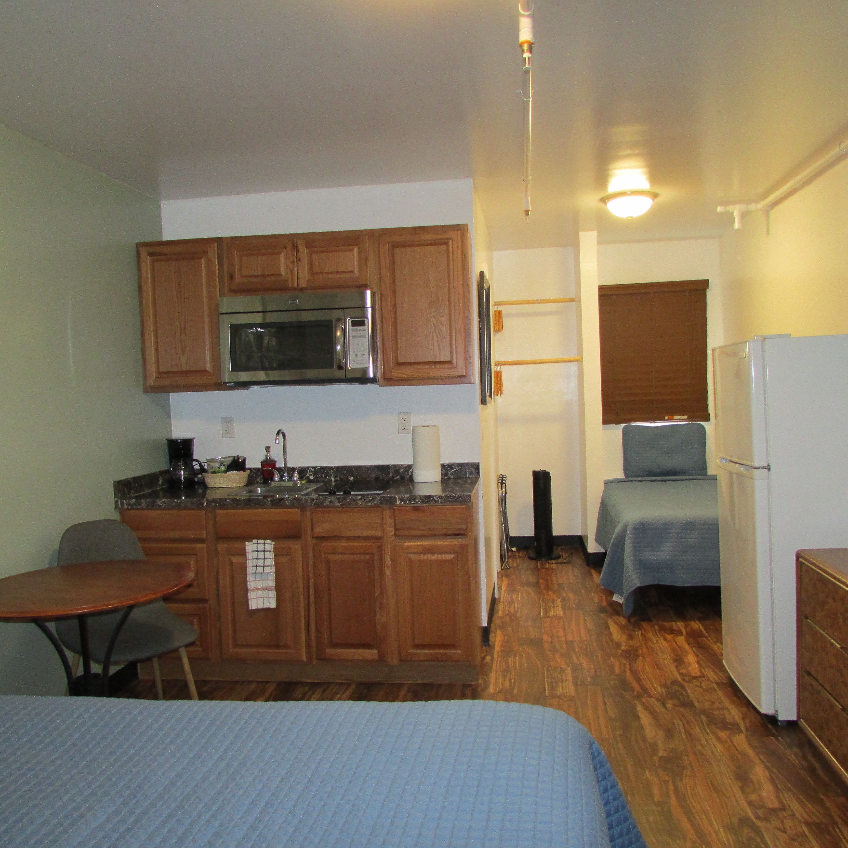 A compact kitchenette with a microwave, sink, and wooden cabinets. There is a small table and a chair, and a bed with a blue blanket.