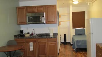 Una kitchenette compacta con microondas, pileta y muebles de madera. Hay una mesita y una silla, y una cama con un edredón azul.