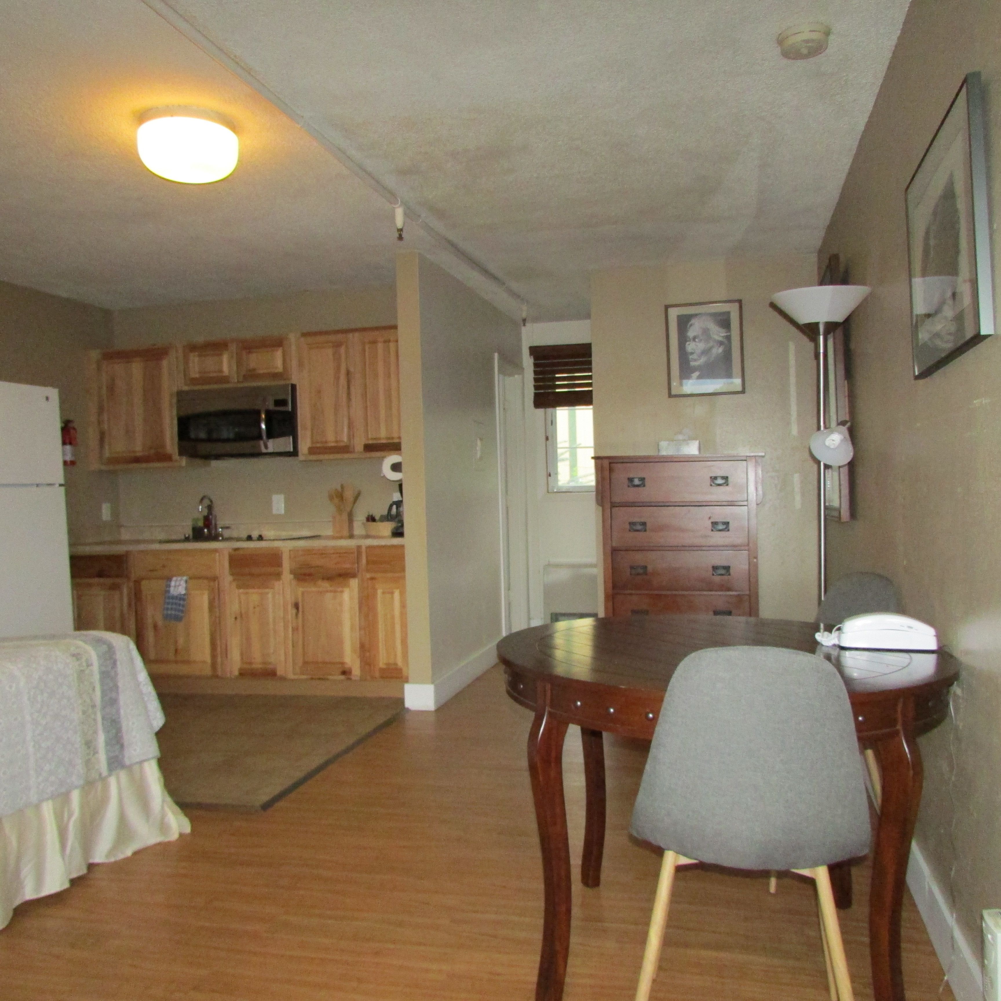 A compact kitchen with wooden cabinets, a microwave, and a refrigerator. There is a dining table with two chairs and a lamp on the wall.