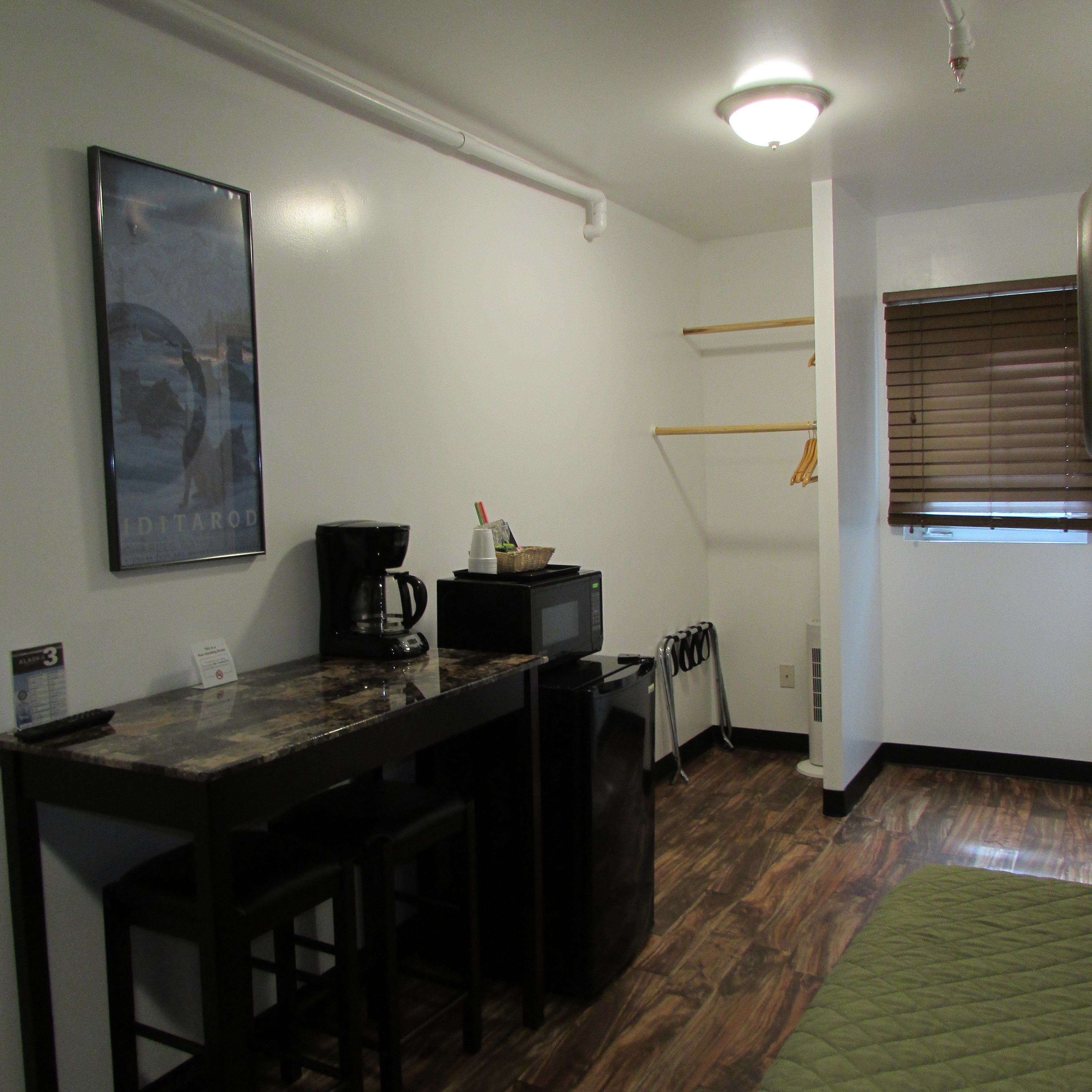 A compact kitchen with a coffee maker, a microwave, and a small refrigerator. There is a wooden bar stool and a green rug on the floor.