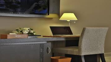 A hotel room with a desk, a laptop, a lamp, a TV mounted on the wall, a chair, and a trash bin.