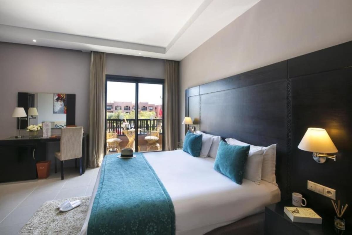 A hotel room with a large bed, a desk, a chair, a balcony with outdoor seating, and a view of buildings.