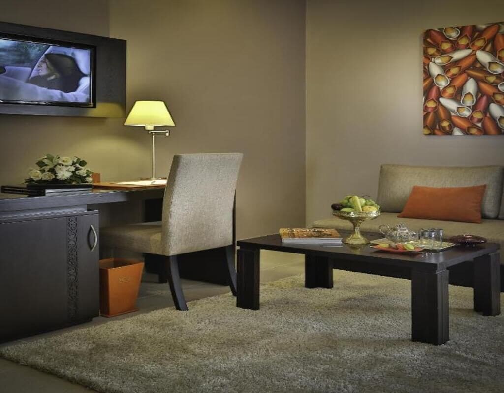 Ein Hotelzimmer mit Flachbildfernseher an der Wand, einem Schreibtisch mit Lampe, einem Sessel, einer Couch mit orangefarbenem Kissen und einem Couchtisch mit Fruchtschale und Buch.