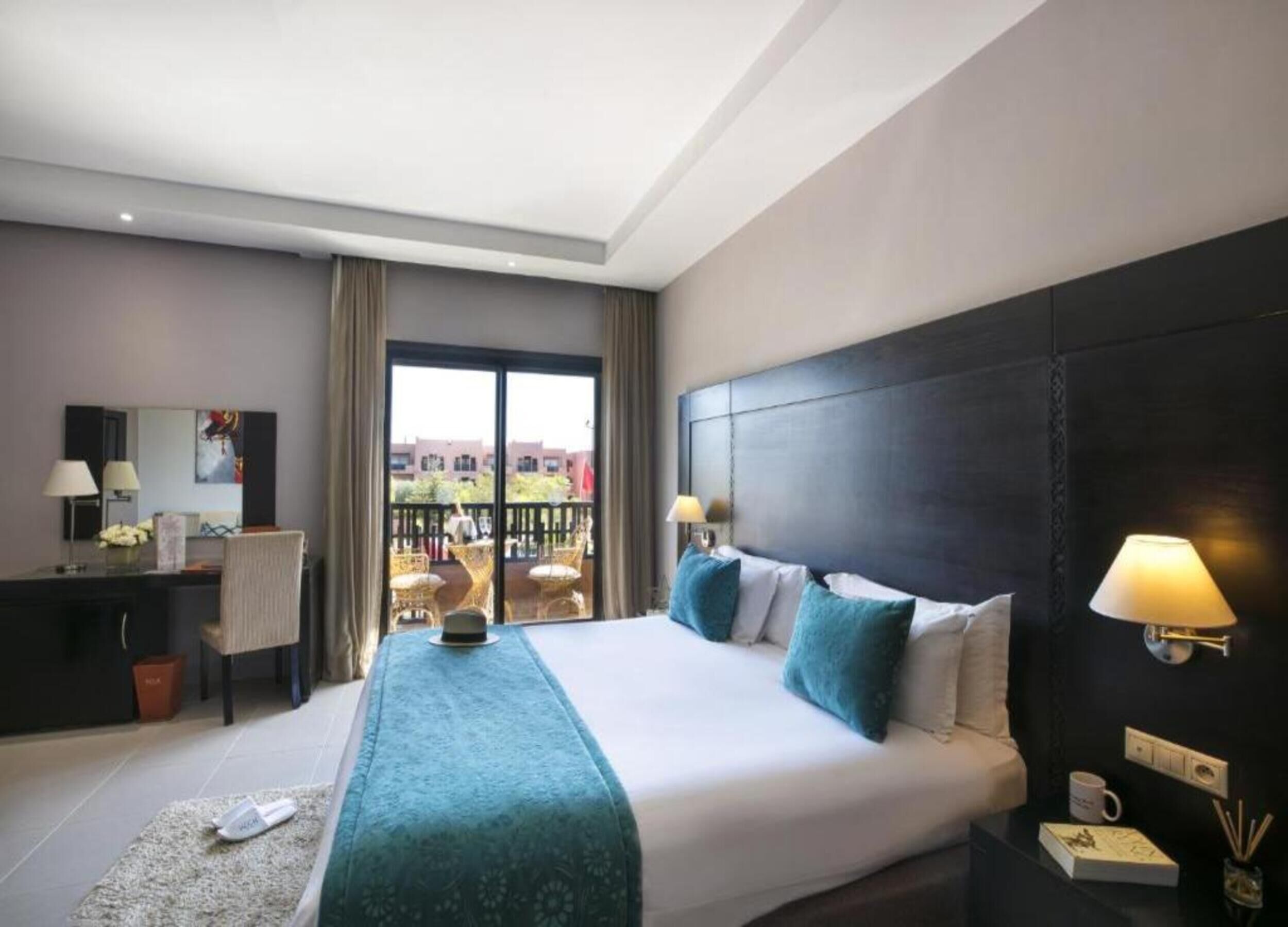 Ein Hotelzimmer mit einem großen Bett, einem Schreibtisch, einem Sessel, einem Balkon mit Sitzgelegenheit und Blick auf Gebäude.