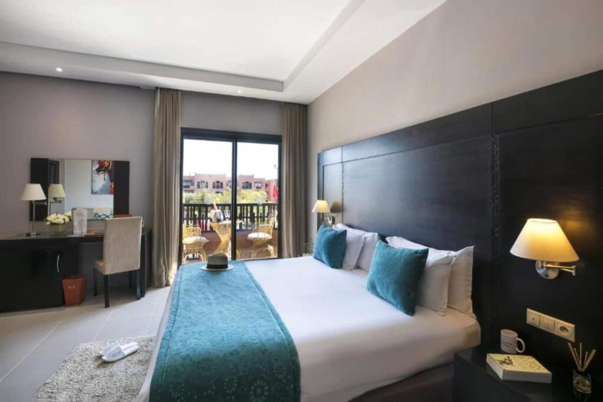 Habitación de hotel con una cama grande, un escritorio, una silla, un balcón con asientos exteriores y vistas a edificios.
