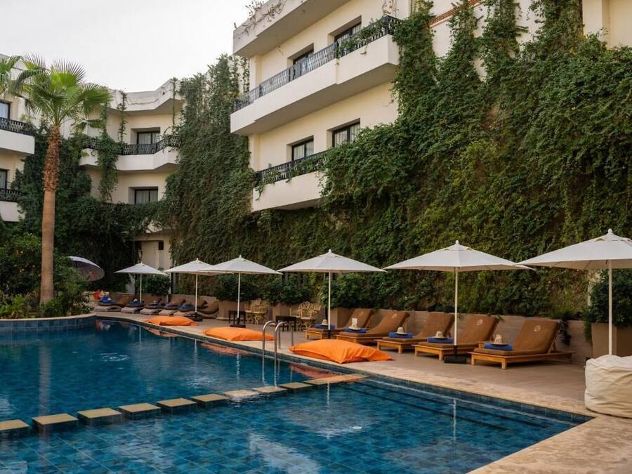 Una piscina con sillas de descanso y sombrillas, rodeada por un edificio de fachada blanca y hiedra verde.
