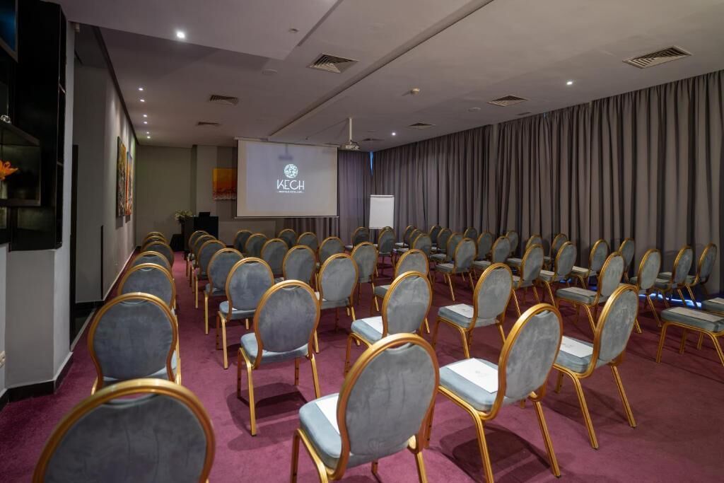 A conference room with rows of grey chairs, a projector screen, and a large screen displaying 'KECH'.