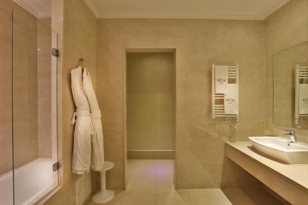 Ein modernes Badezimmer mit beigen Fliesen, einer Glasdusche, einer weißen Badewanne, einem Waschbecken inklusive Spiegel und einem Handtuchhalter mit Bademantel.