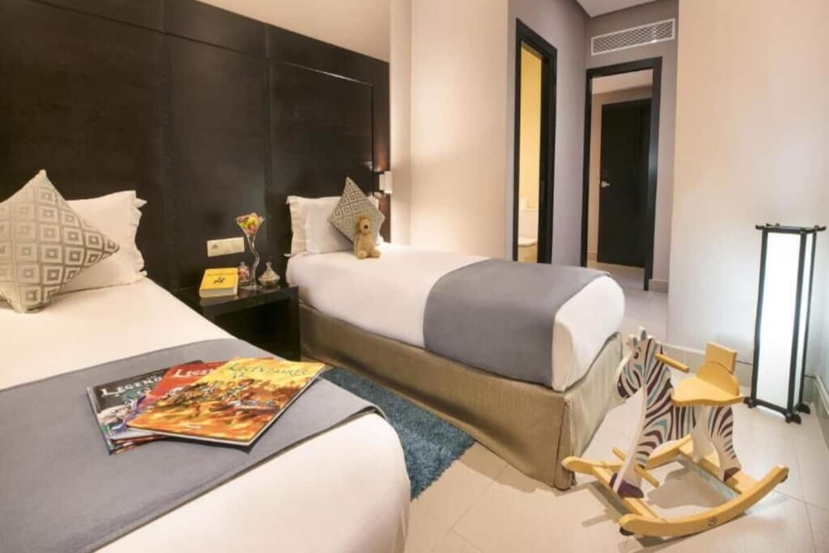Una habitación de hotel con dos camas, un sillón mecedor estampado con diseño de cebra, una revista sobre la cama y una mesita con una lámpara.