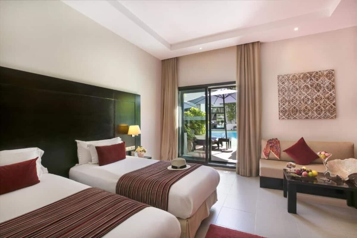 Habitación de hotel con dos camas, un sofá, una mesa de comedor y vistas a una piscina y zona exterior.