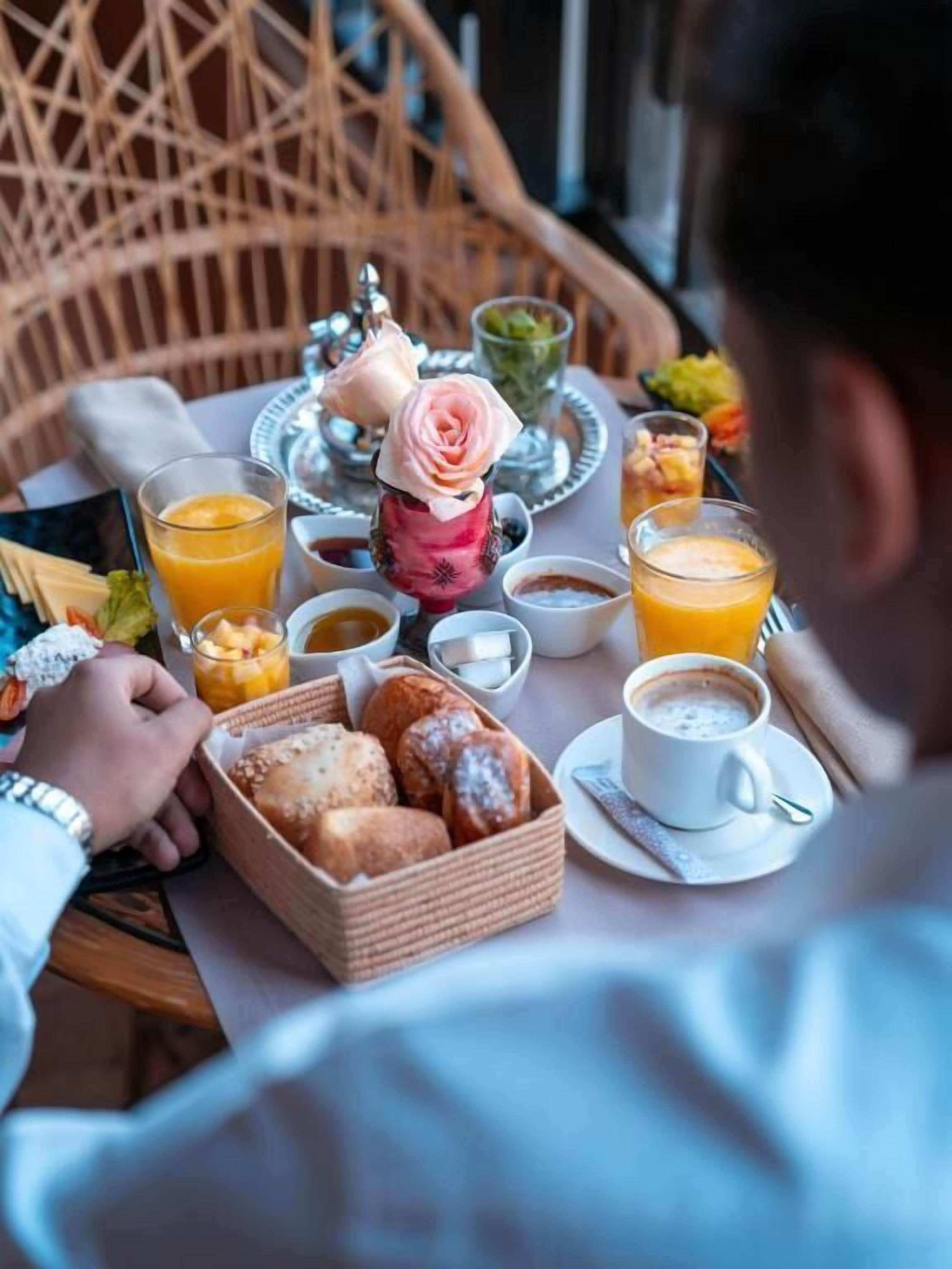 Un desayuno con una silla de mimbre, una cesta de pasteles y una mesa con diversas opciones para el desayuno, incluyendo jugo de naranja y café.