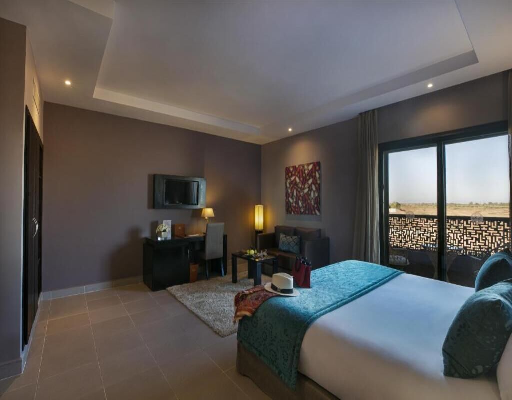 A modern hotel room with a large bed, a desk, a chair, a TV, and a view of a beach.