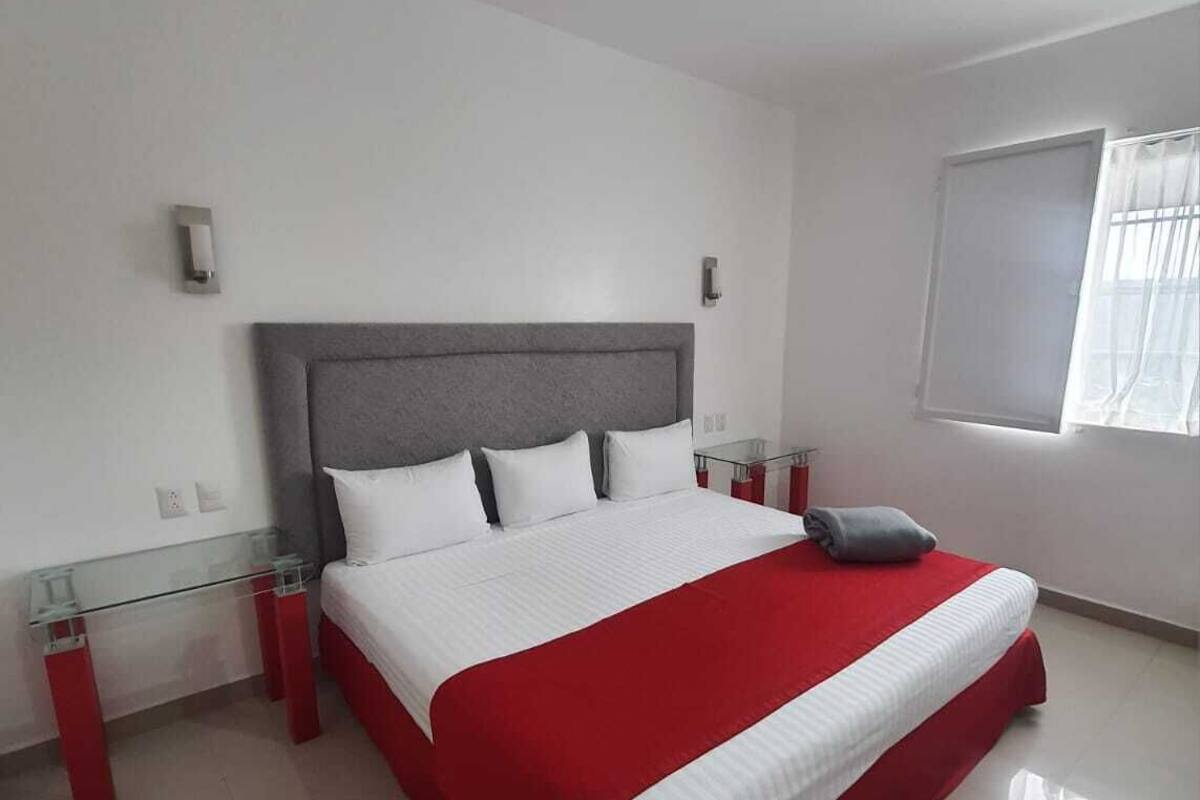 A modern bedroom with a large bed, a glass bedside table, and a window with curtains.