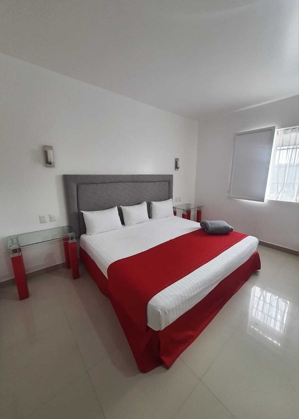 Un dormitorio moderno con una cama grande, una mesita de noche de vidrio y una ventana con cortinas.
