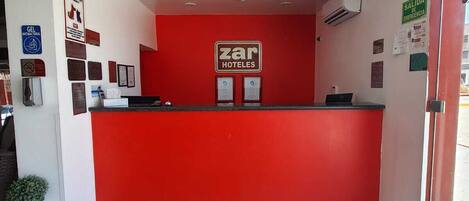 Zona de recepción de hotel con un mostrador rojo, un cartel que dice "Zar Hoteles" y un letrero de "SALIDA DE EMERGENCIA".