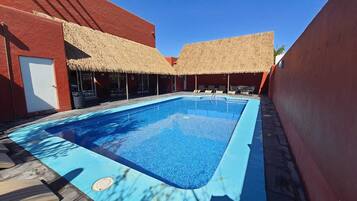 Piscina com borda azul, rodeada por edifícios vermelhos e estruturas com telhados de palha.