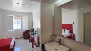 Una habitación de hotel moderna con una cama grande, un escritorio con silla, un baño con lavamanos y espejo, y un ventanal que proporciona luz natural.