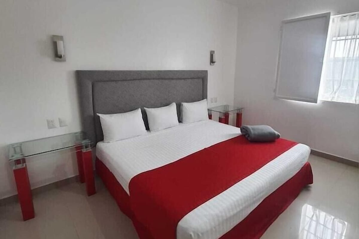 A modern hotel room with a large bed, a glass table, and a window with curtains.