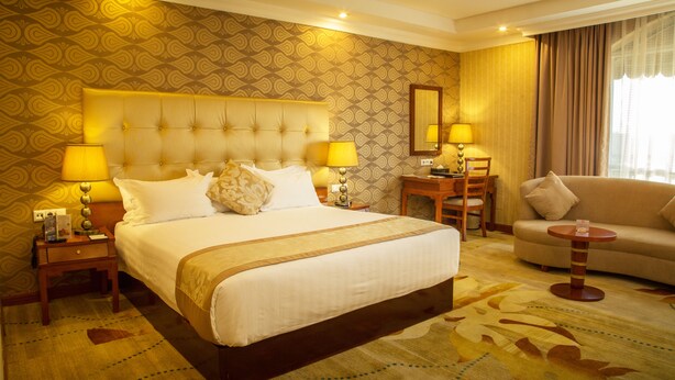 A hotel room with a large bed, two bedside tables with lamps, a small table with a chair, and a sofa. - Jupiter International Hotel Bole (Addis Ababa)