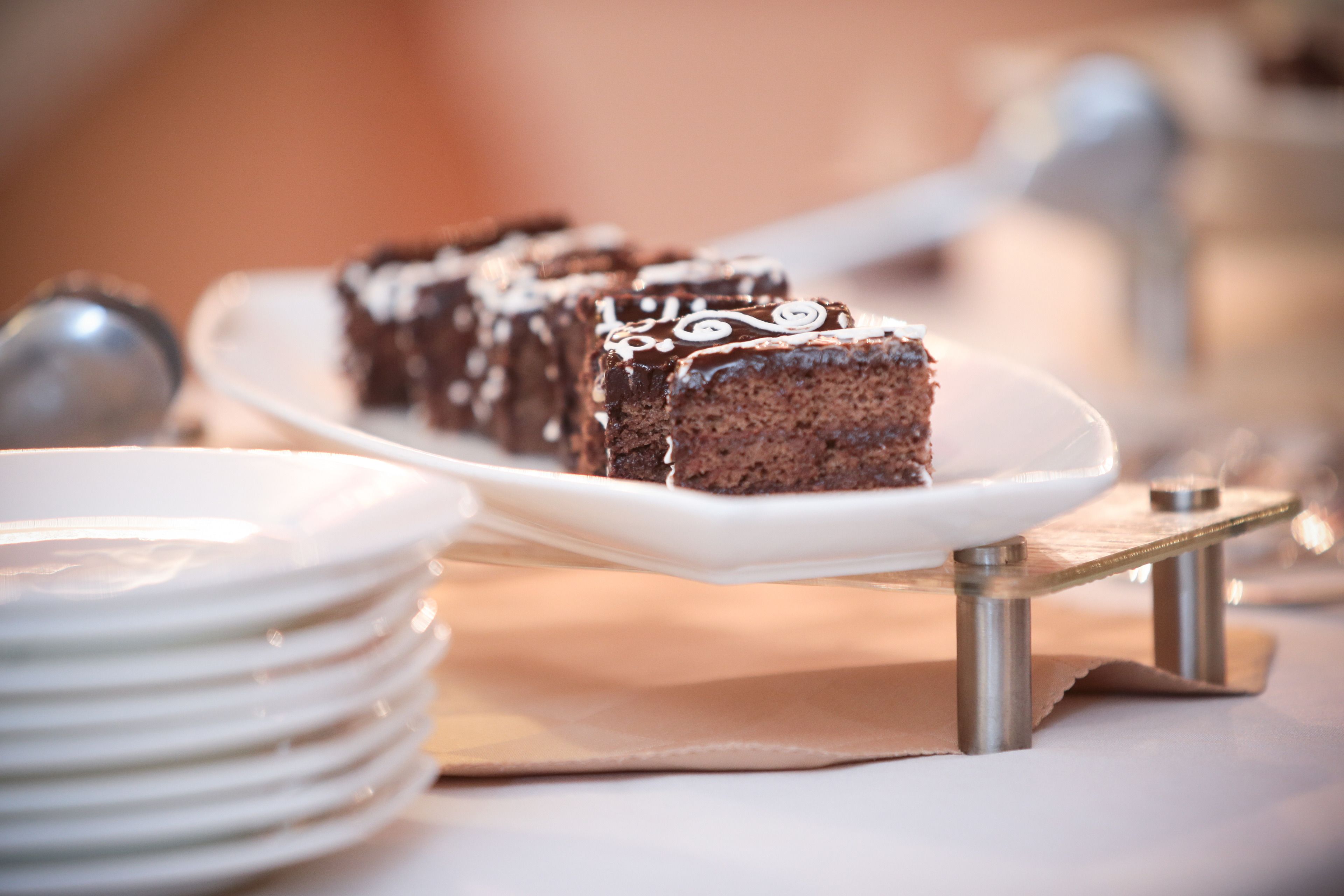 A dessert tray with chocolate cake slices on a white plate, accompanied by stacked white plates and a metal stand.