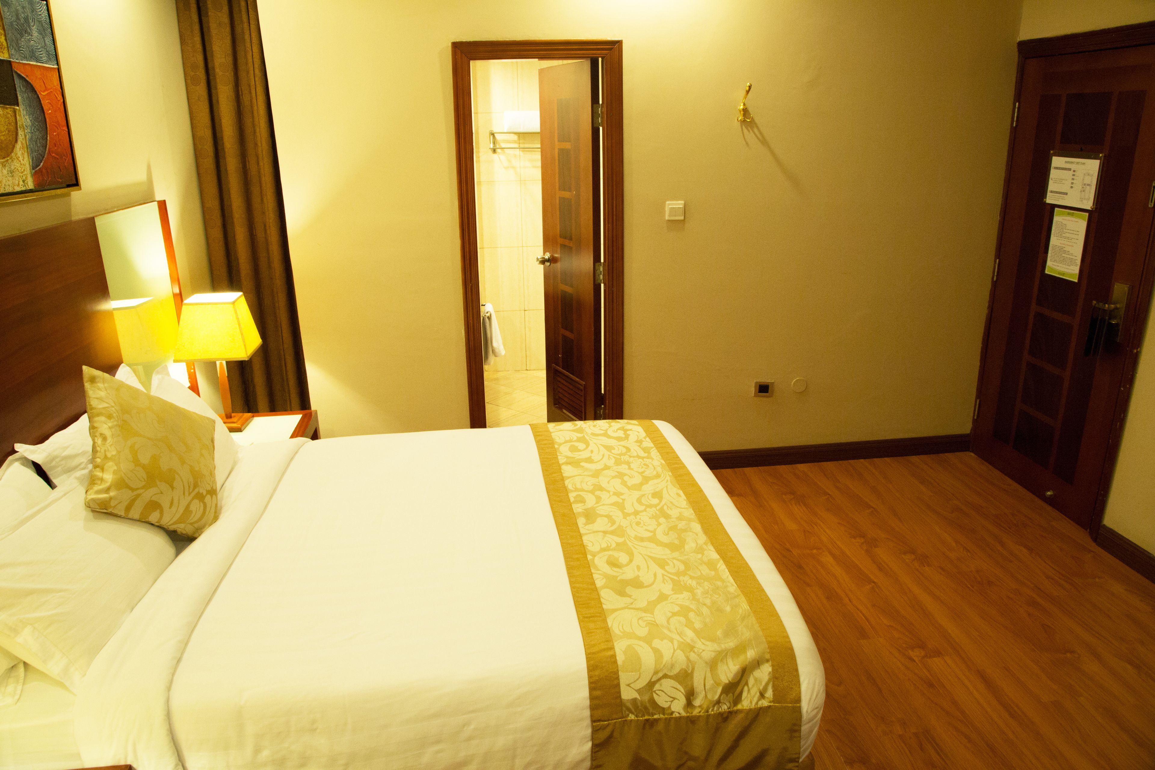 Une chambre d’hôtel avec un lit, une table de chevet avec une lampe, une porte en bois et une salle de bain visible à travers une porte ouverte.