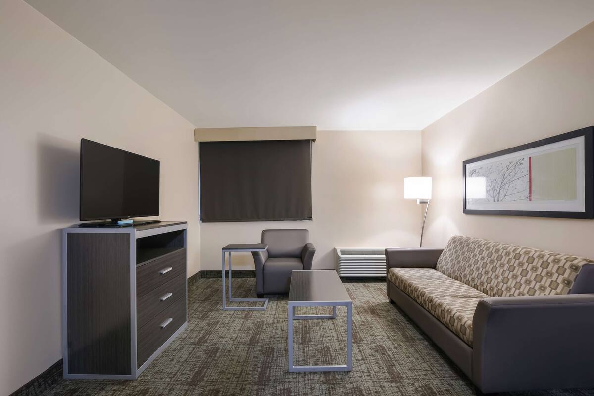 Ein Hotelzimmer mit Flachbildfernseher auf einem Schrank, einer Couch, einem Sessel, einem kleinen Tisch und einer Wandlampe.