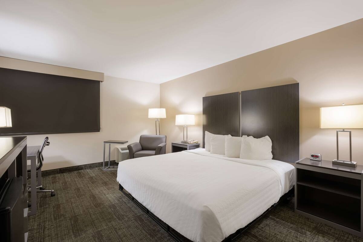 Ein Hotelzimmer mit einem großen Bett, zwei Nachttischen mit Lampen, einem Schreibtisch mit Stuhl und einem an der Wand befestigten Fernseher.