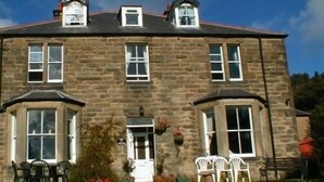 A stone building with a central entrance, two side wings, and a small porch area with chairs. - The Haven (Morpeth)