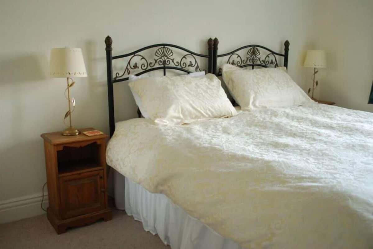 A neatly made bed with a white bedspread and pillows, a wooden nightstand with a lamp, and a metal headboard.