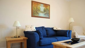 A living room with a blue sofa, two wooden side tables, a painting on the wall, and a lamp on each table. - The Haven (Morpeth)