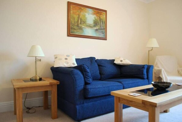 A living room with a blue sofa, two wooden side tables, a painting on the wall, and a lamp on each table. - The Haven (Morpeth)